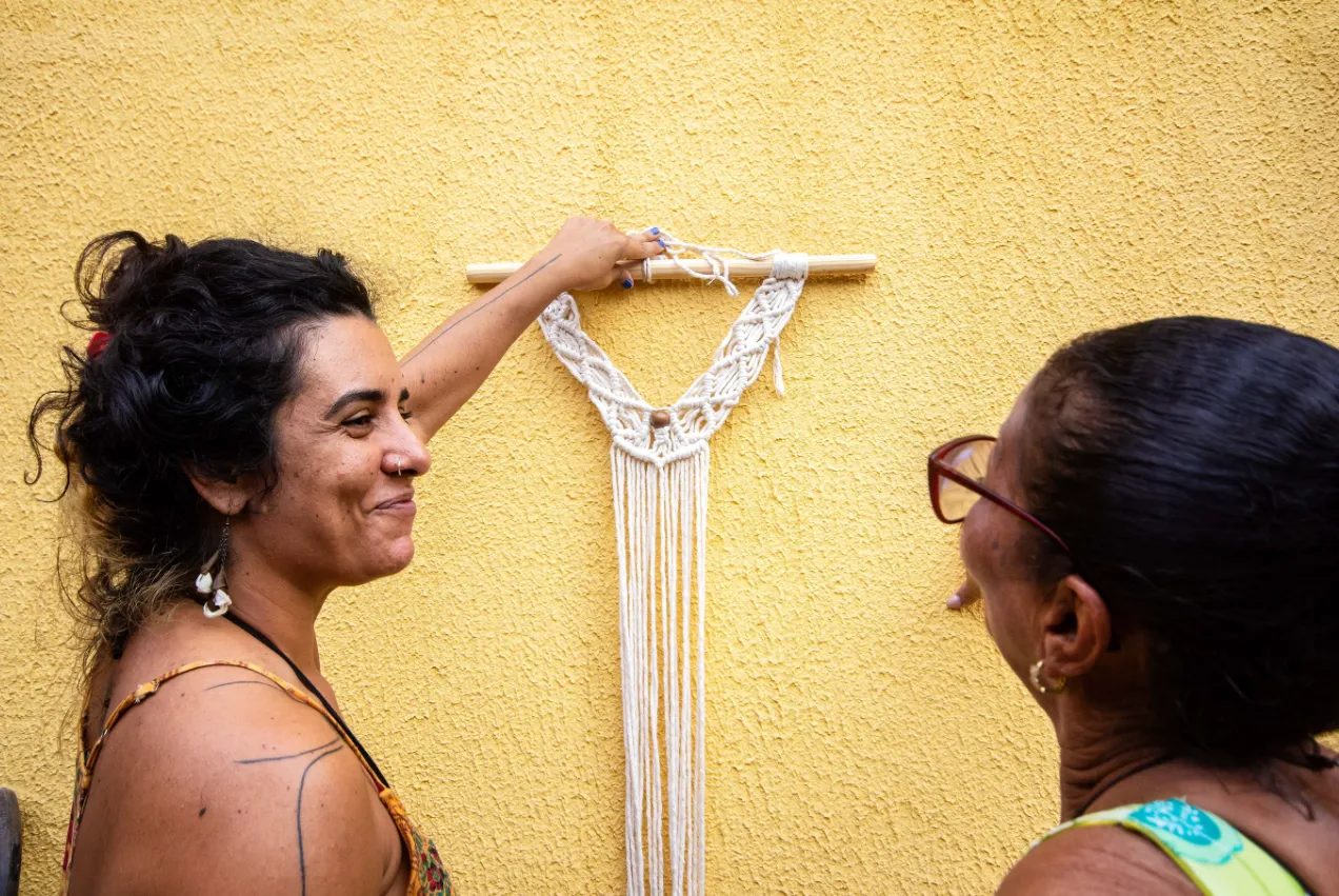 Duas mulheres admiram um painel de macramê numa parede amarela